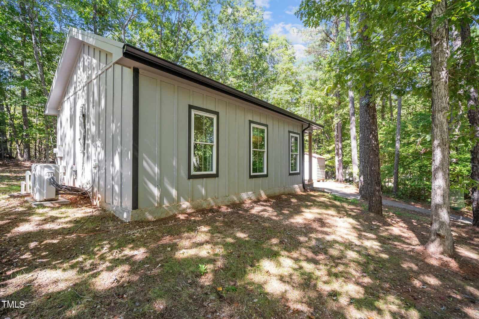 A small, modern light-gray house with vertical siding and multiple windows sits in a wooded area with tall trees and dappled sunlight on the ground.
