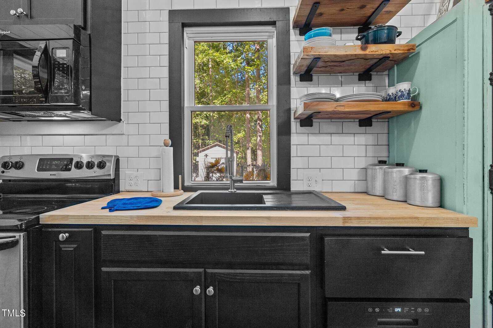 Modern kitchen with black cabinets, a wood countertop, a black sink, open shelves with dishes, white subway tile backsplash, and a window with a view of trees and a backyard shed.