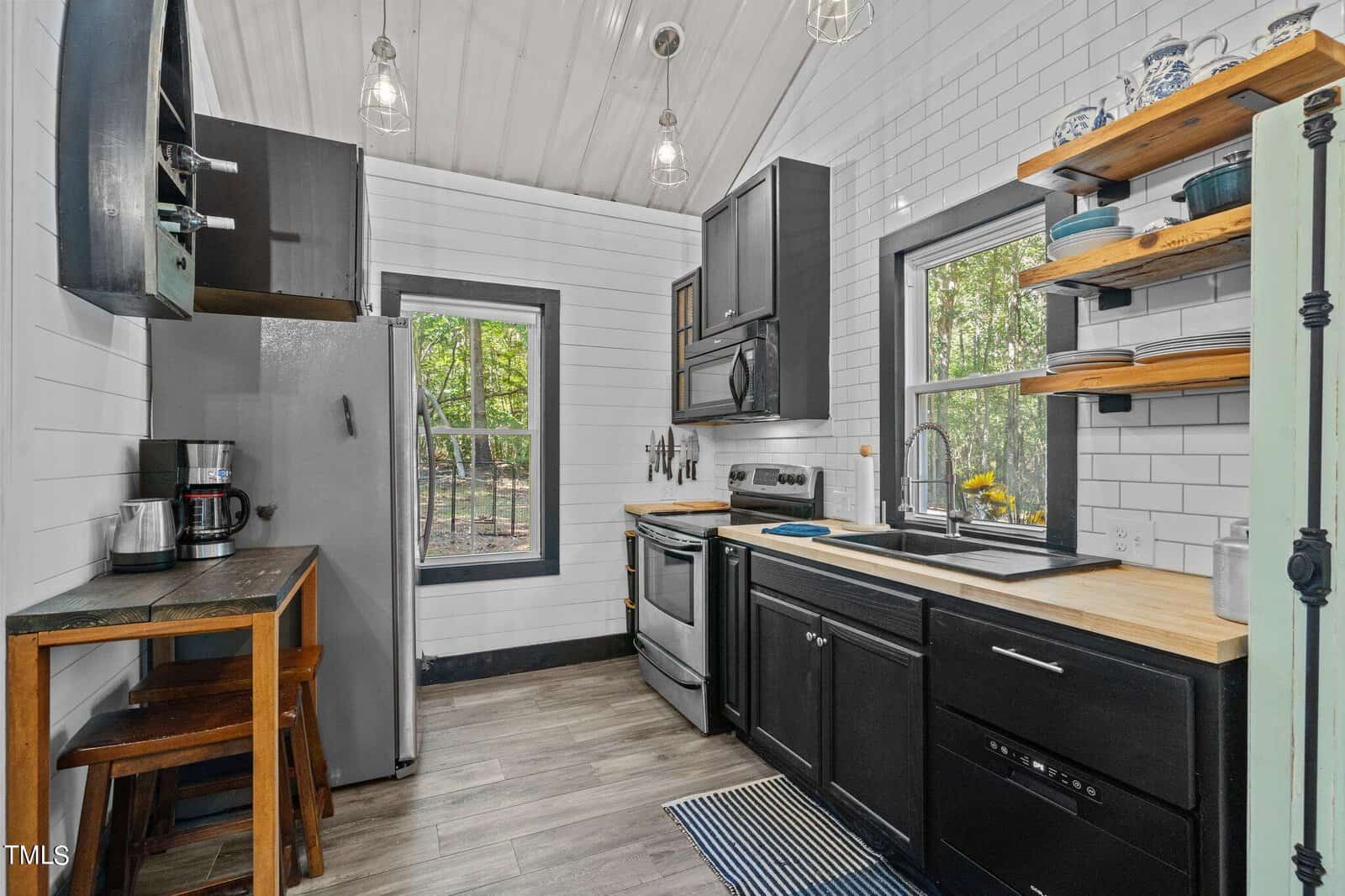 Modern kitchen with white subway tile walls, black cabinets, stainless steel appliances, open wooden shelves, a window over the sink, a small table with stools, and wood-look flooring. Bright, natural light from big windows.