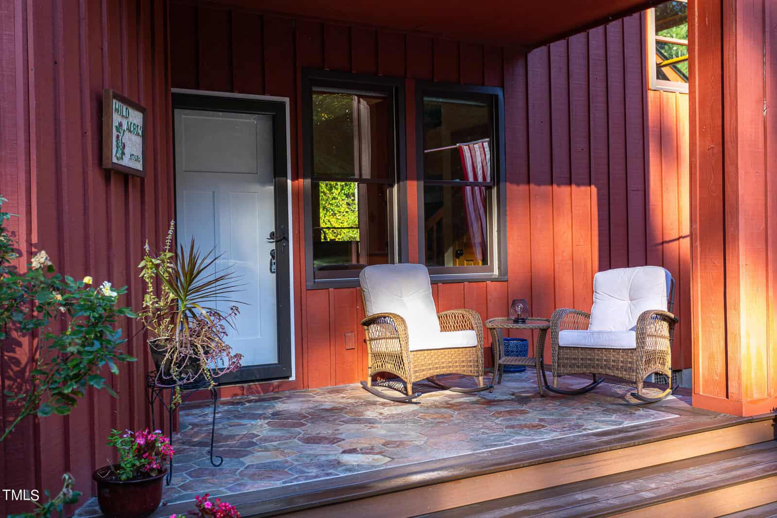 Cozy porch with two wicker chairs and white cushions on a stone-tile floor. A small side table holds a glass, and potted plants decorate the space. The red wooden siding gives a warm, inviting appearance, enhanced by the soft sunlight.