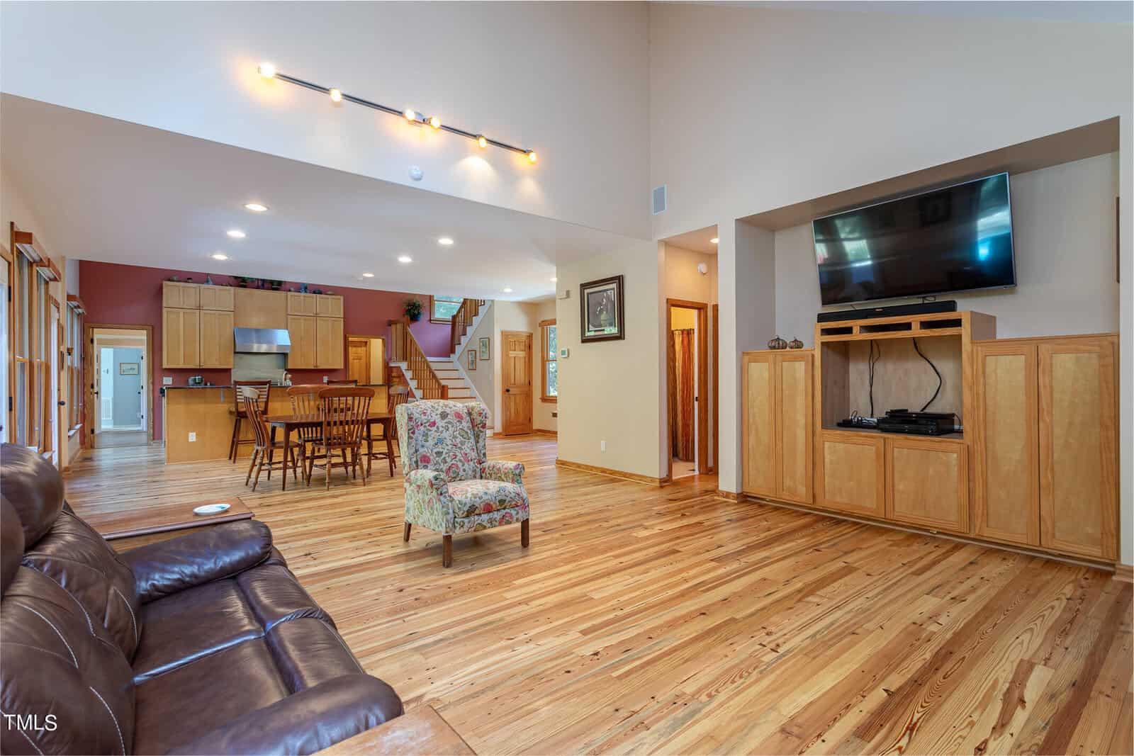 Spacious living area with wooden floors and high ceilings. A brown leather sofa and a floral armchair face a large wall-mounted TV above a wooden cabinet. The open-plan kitchen with wooden cabinetry and a dining table is visible in the background.