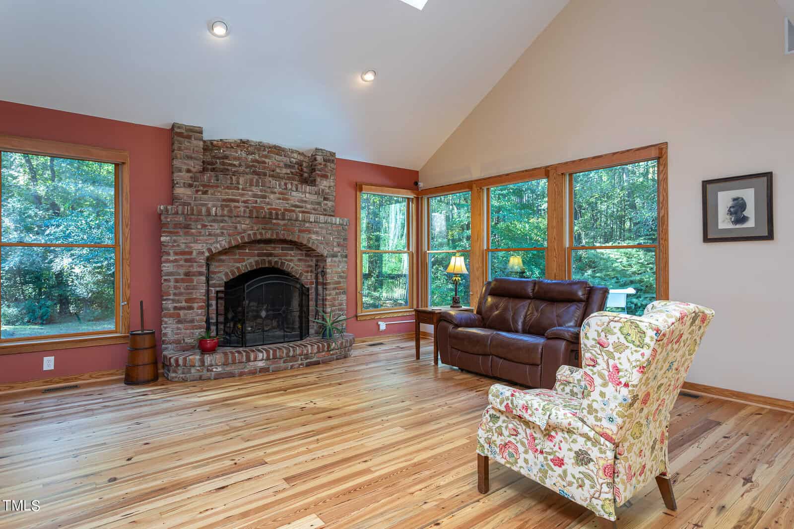 A cozy living room with large windows showcasing a forest view. There's a brick fireplace, a brown leather sofa, a floral armchair, and wood flooring. Warm, earth-toned walls and a framed picture complete the setting.