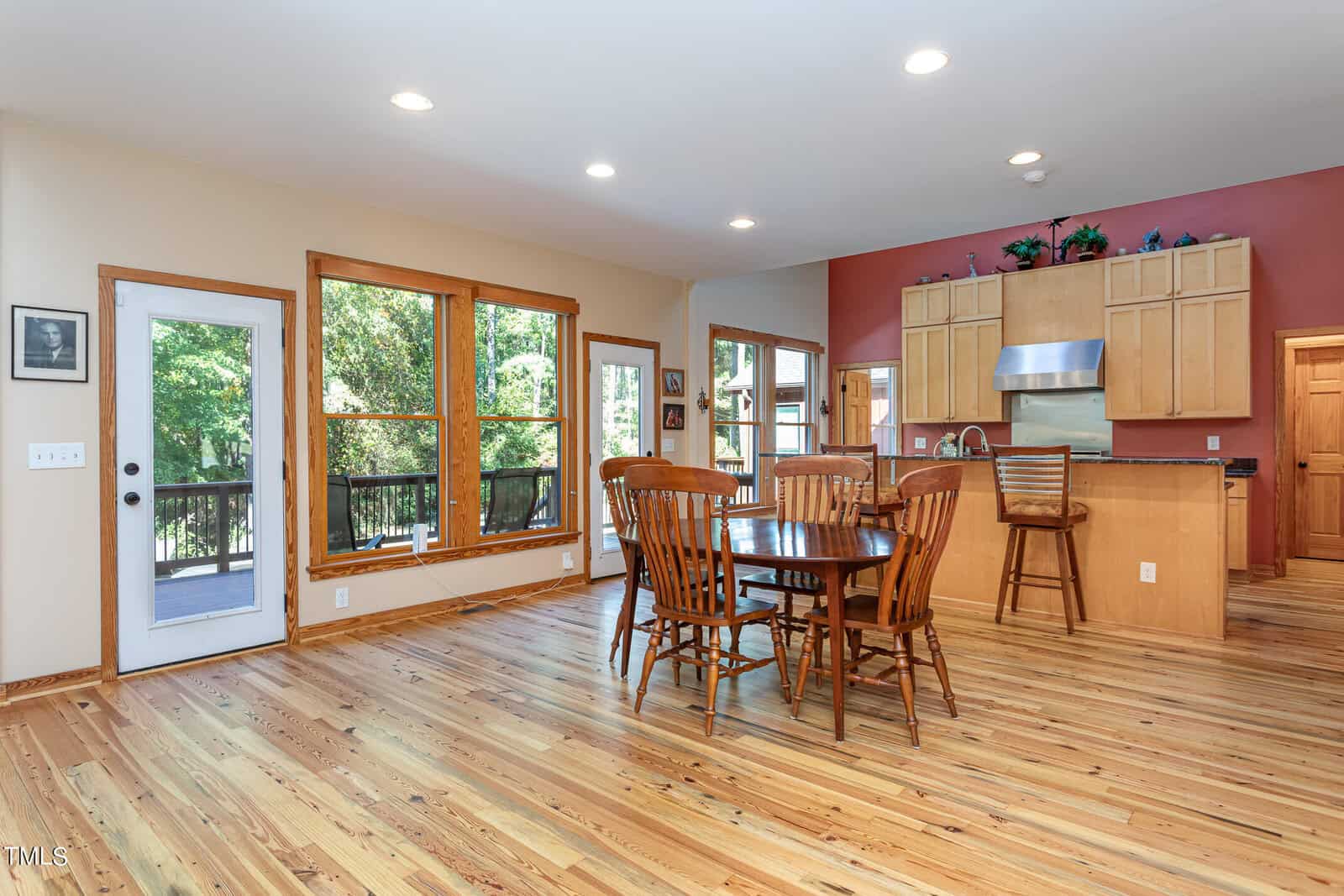 A spacious kitchen and dining area with wooden floors and cabinets. There is a round dining table with chairs in the center. Large windows and a glass door open to a deck, letting in natural light and offering a view of trees outside.