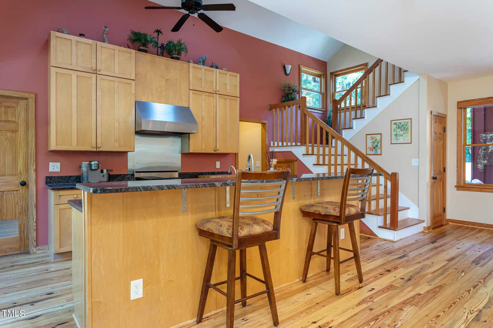 A spacious kitchen with light wood cabinets and a center island with two wooden bar stools. The room features a red accent wall, stainless steel appliances, and a staircase leading to the upper level, with plants and framed art as decor.