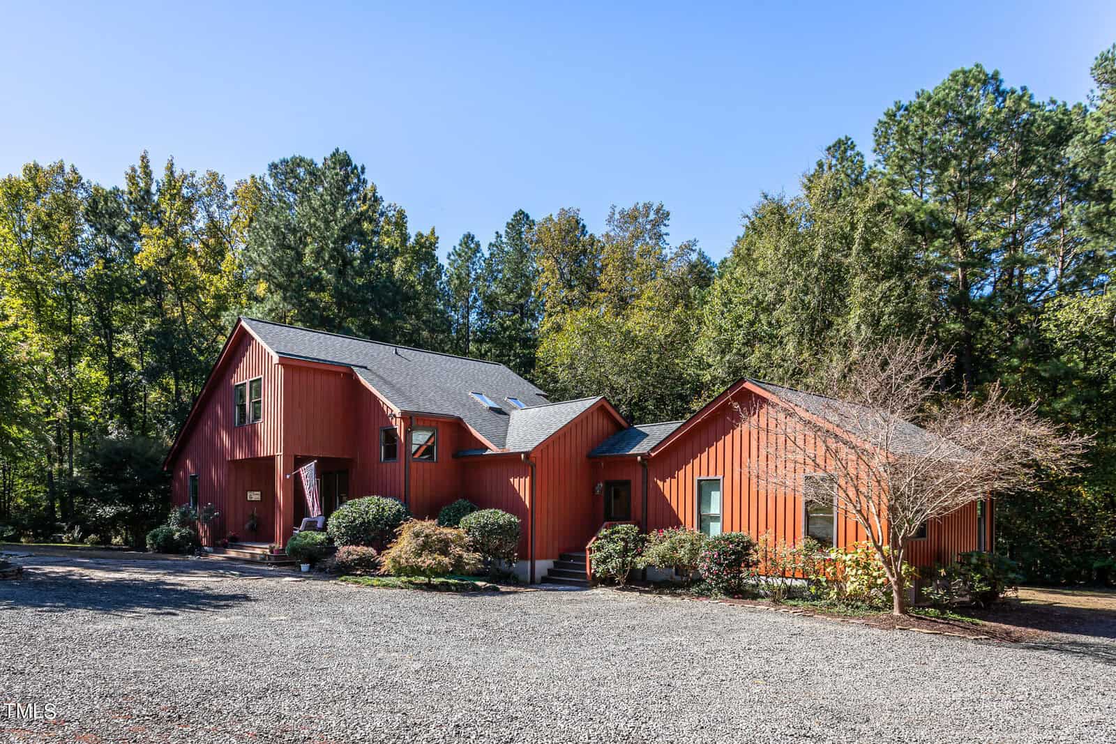 A modern red wooden house with a gray roof is surrounded by trees. The front yard features shrubbery and a gravel driveway under a clear blue sky.