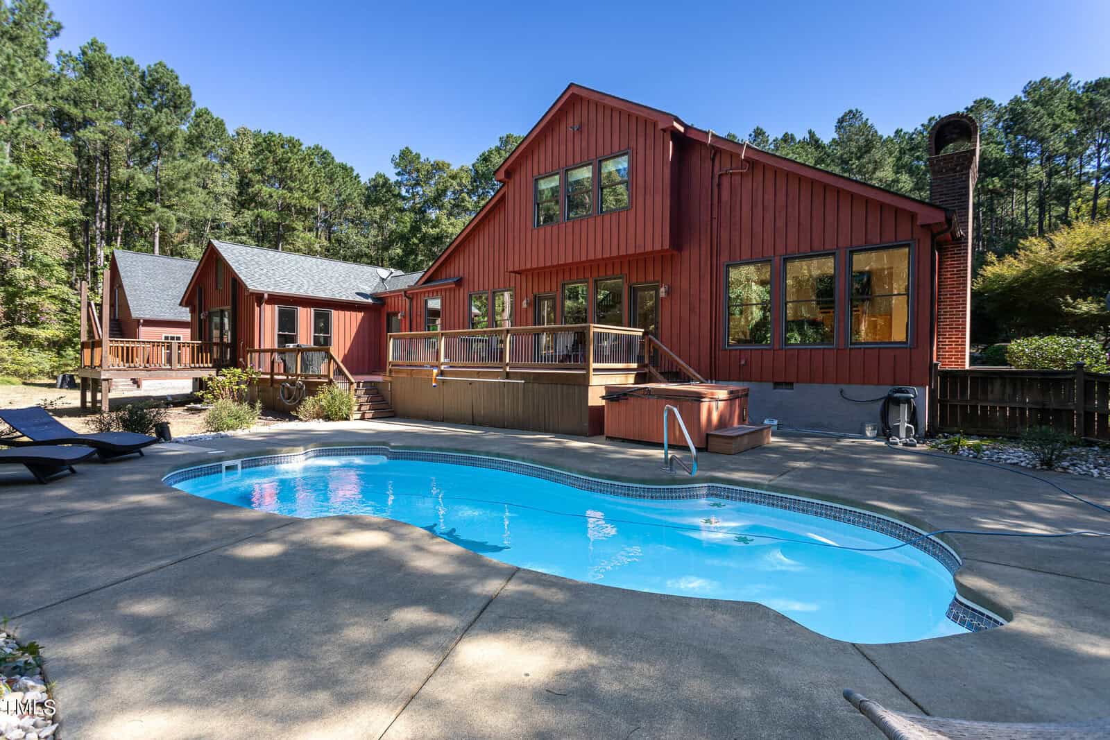 Nestled in a wooded area at 1141 Fleming Road, Creedmoor, a red wooden house with large windows and a charming chimney overlooks a swimming pool. The concrete patio surrounds the pool, which sits next to a smaller wooden building, creating an inviting retreat.