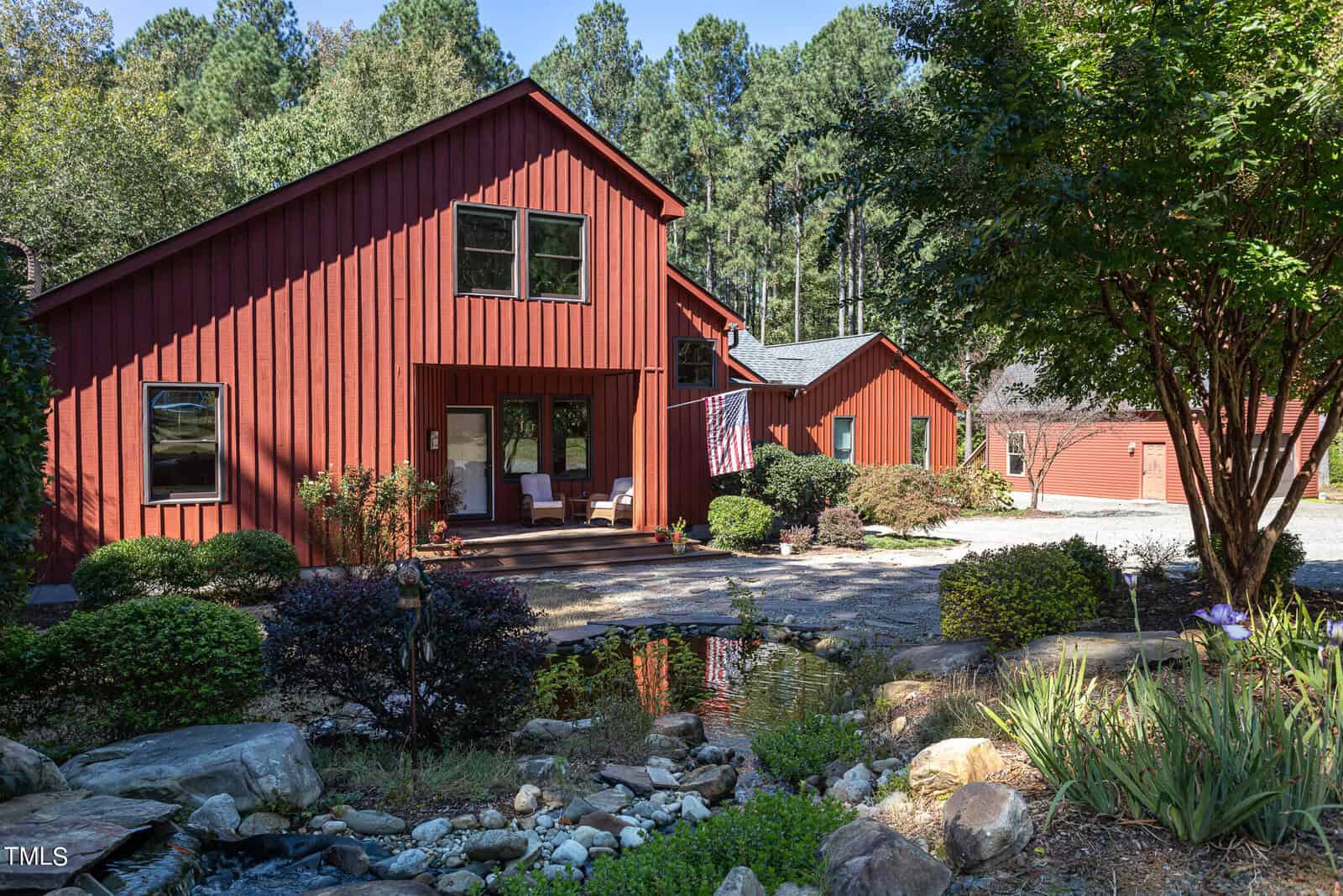 A red wooden house with large windows is surrounded by lush greenery. A small pond with rocks is in the foreground, and a tree adds shade. Clear blue sky is visible in the background.