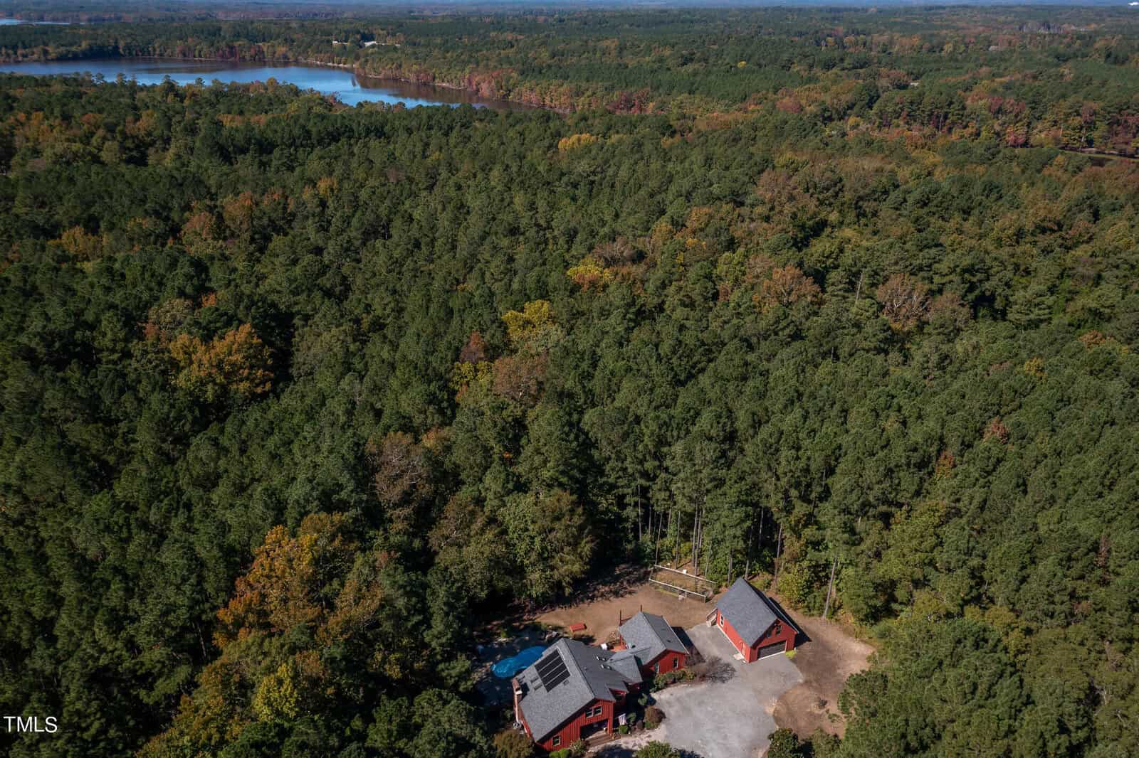 Aerial view of a dense forest with a red-roofed house surrounded by trees. A dirt driveway leads to the house. In the background, a large lake is visible amidst the greenery.