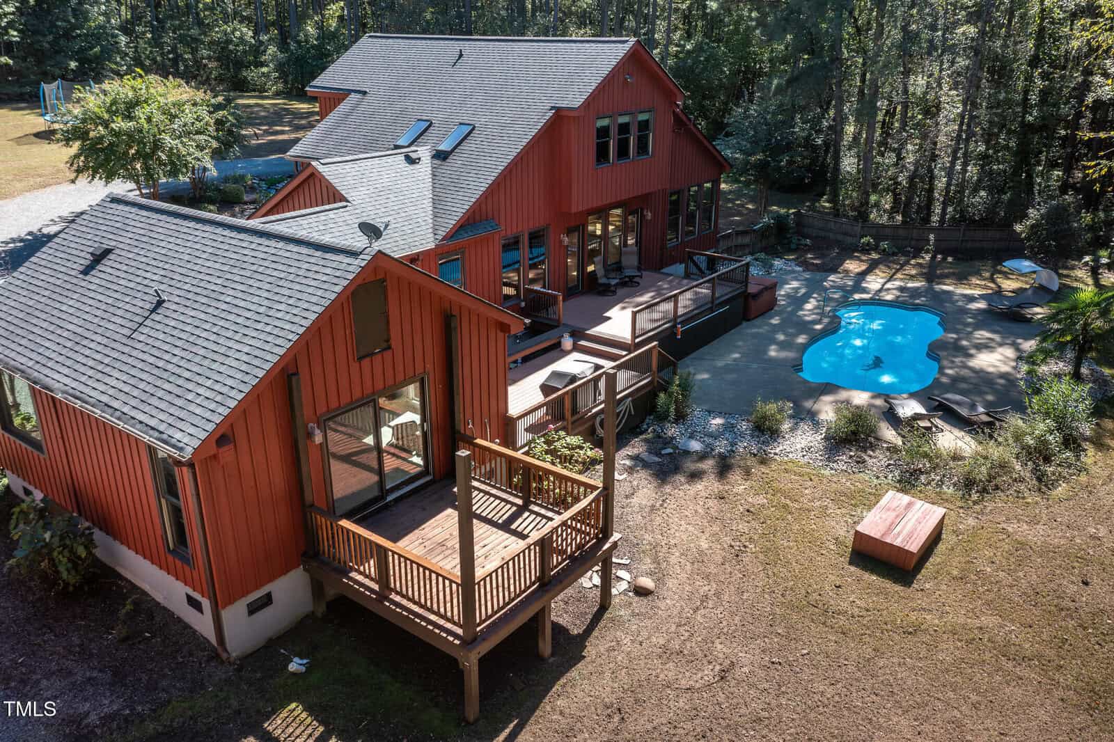 Aerial view of a large red house with a sloped roof surrounded by trees. It features multiple decks and a backyard pool with a curvy shape. There is a small shed near the house and a driveway in the background.
