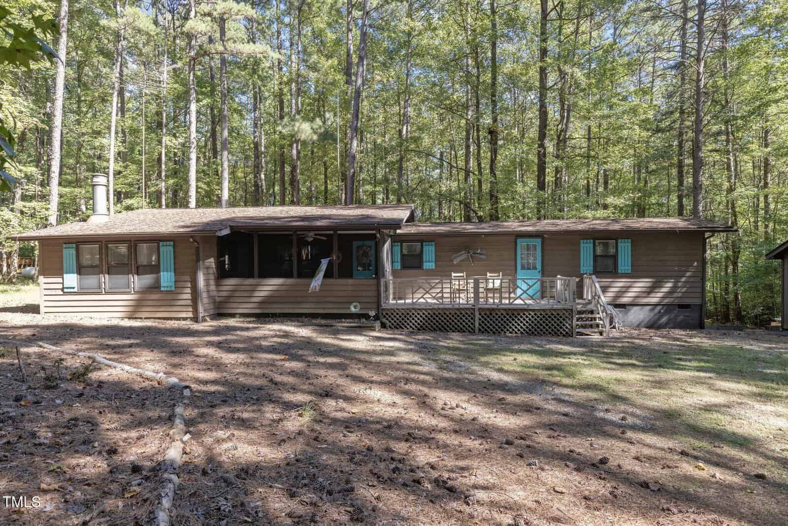 Nestled on 396 Forest Home Lane in Henderson, this rustic wooden cabin is surrounded by tall trees. With teal shutters and doors, a chimney, and a cozy deck with patio chairs, the sun filters through the forest, casting a serene atmosphere.