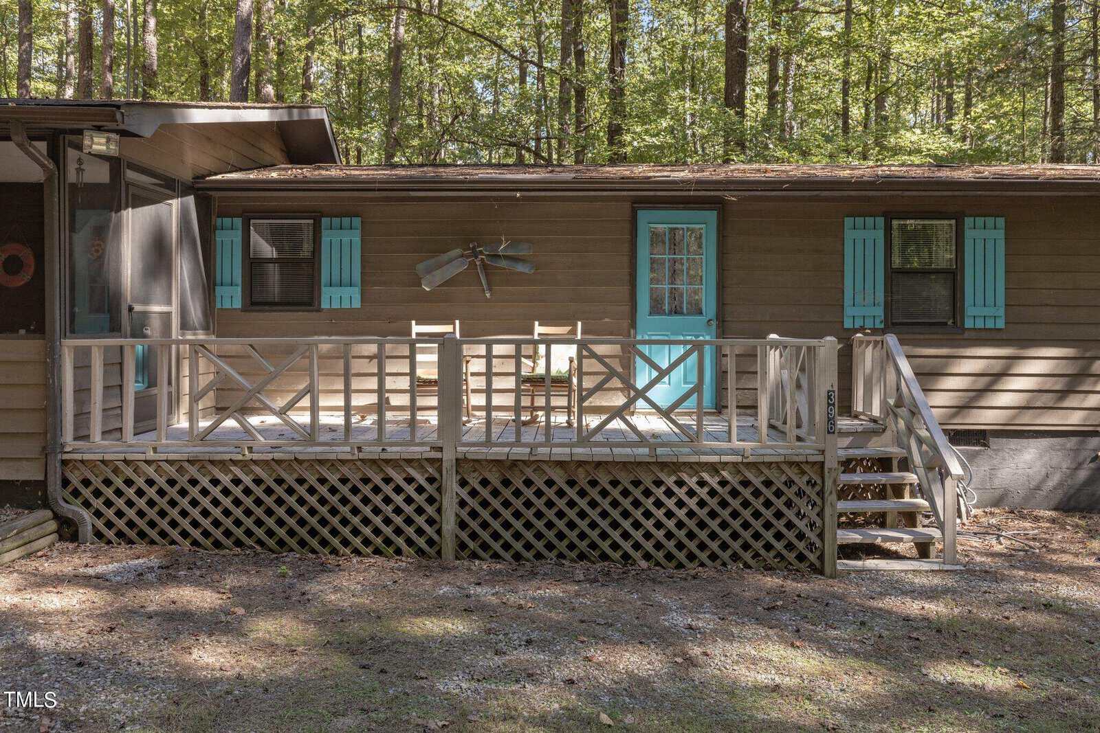 A rustic cabin with a porch in a wooded area. The porch has two wooden chairs and is accessible by a small set of stairs. The cabin has blue window shutters and a teal door. Surrounded by tall trees, the cabin has a peaceful, natural setting.