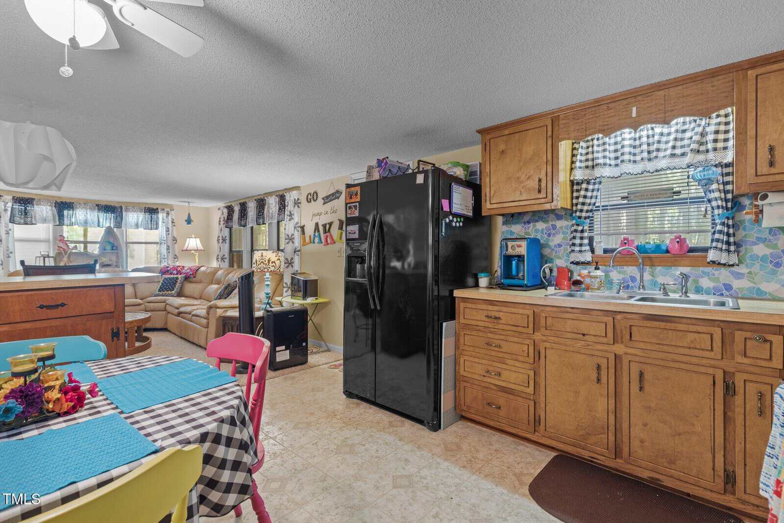 A cozy kitchen with wooden cabinets, a black fridge, and a sink. A dining table with a colorful striped tablecloth is in the foreground, with painted chairs. The room is bright, with a sofa and window in the background.