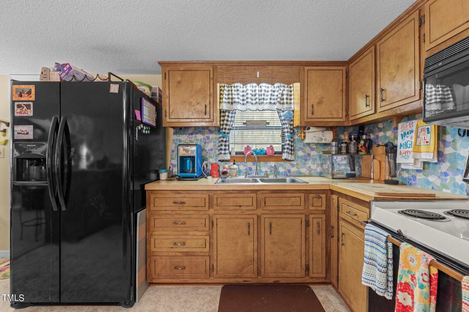 A cozy kitchen with wooden cabinets, a black refrigerator, and an electric stove. A colorful backsplash adds vibrancy, and there are various items on the countertops, including a blue coffee maker. A checkered curtain hangs above the sink.