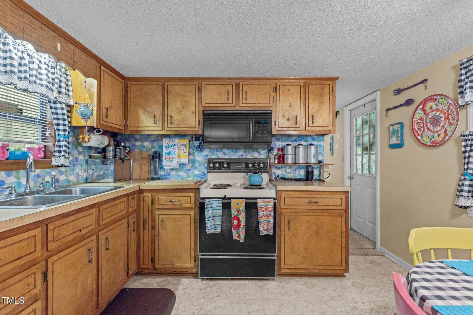 A cozy kitchen features wooden cabinets, a stove with colorful towels, and a blue kettle. Playful decor includes bright plates, utensil art on the walls, and cheerful checkered curtains above the sink. A door with a window is visible in the corner.