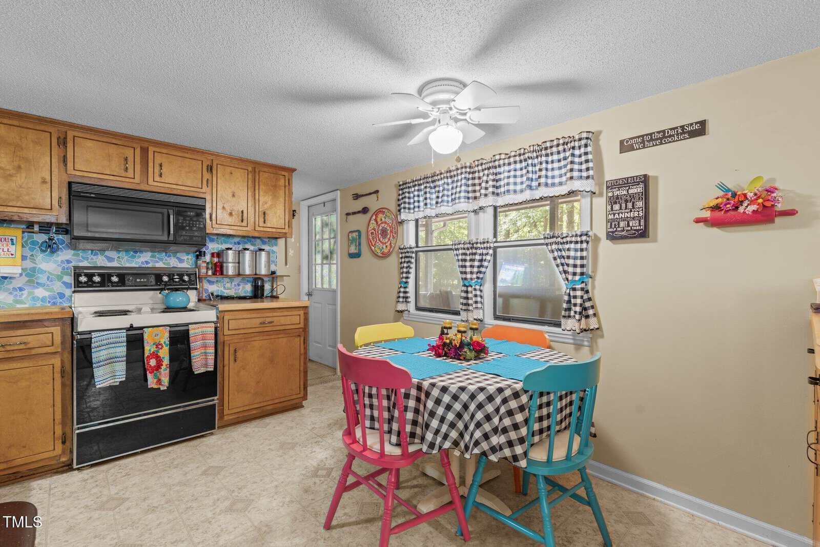 A cozy kitchen with wooden cabinets, a black stove, and a colorful backsplash. A table with mismatched chairs and a checkered tablecloth sits under a ceiling fan. The window has checkered curtains, and decorative items adorn the walls.