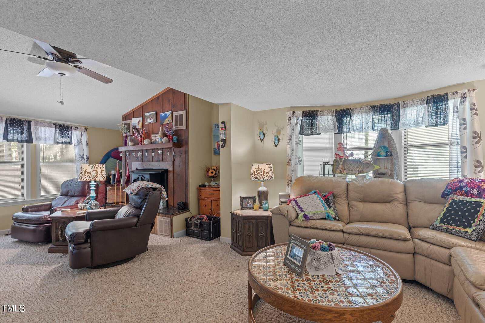 Spacious living room with beige sofas and colorful pillows. A wooden coffee table with decorative bowls sits in the center. To the left are reclining chairs near a brick fireplace, surrounded by wooden shelving with family photos and decor.