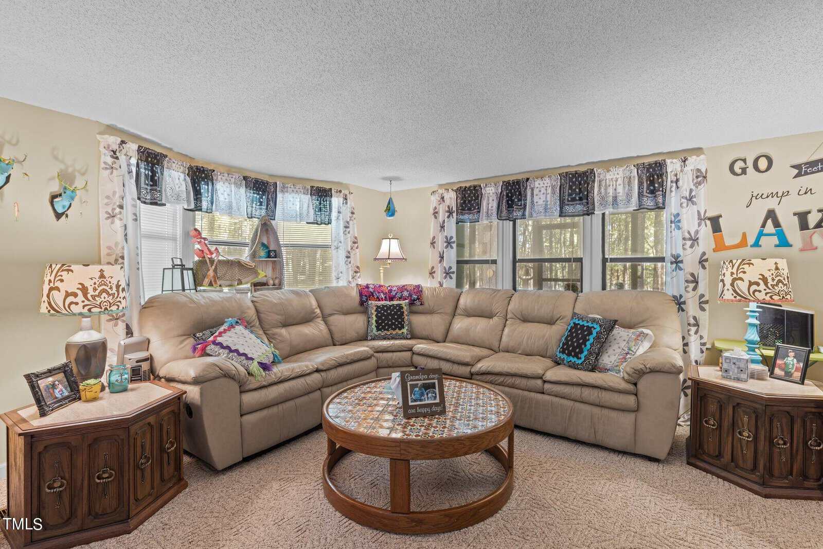 A cozy living room with a large beige sectional sofa and colorful pillows. A round wooden coffee table is in the center, and there are decorated side tables with lamps and picture frames. The walls have nature-themed decor and large windows allow in natural light.