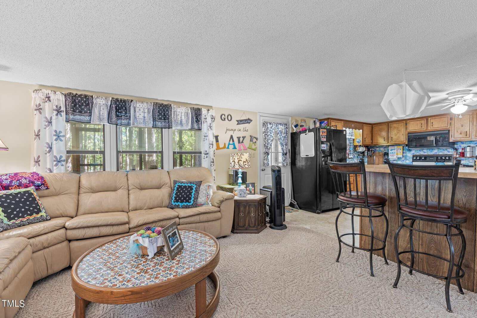 A cozy living room with a beige sectional sofa and patterned cushions faces a wooden coffee table. The kitchen features wooden cabinets and a black fridge. A "Go jump in the lake" sign is on the wall, and large windows let in natural light.