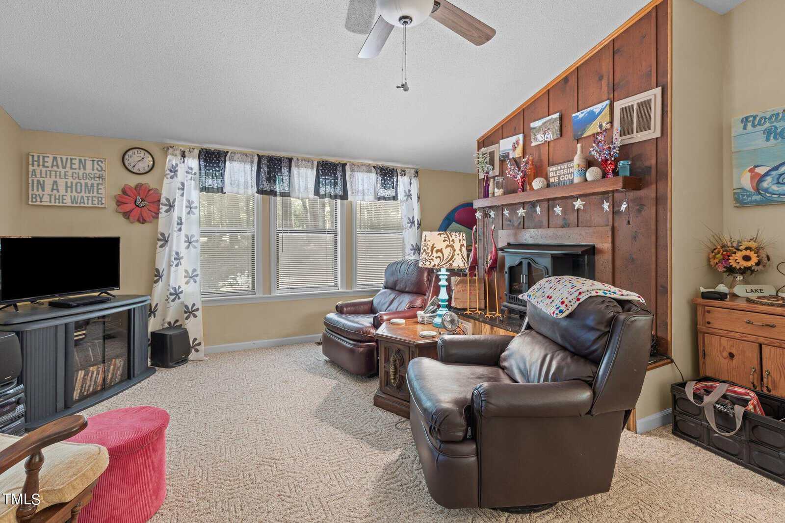 A cozy living room with light carpeting and brown leather armchairs. A fireplace is adorned with decorative items. Large windows with floral curtains allow natural light in. A ceiling fan hangs above, and a TV is placed on a stand in the corner.