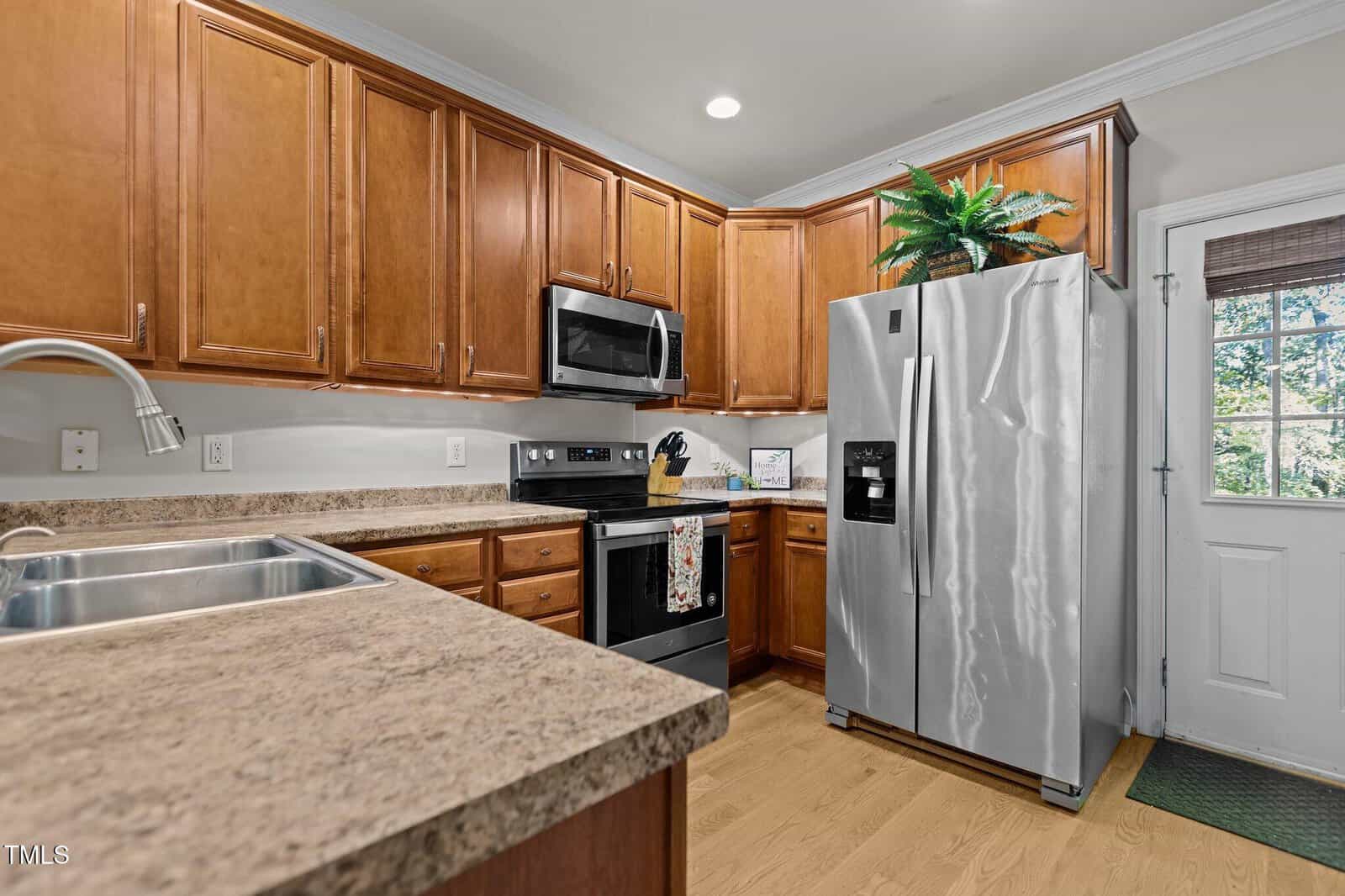 A kitchen with wooden cabinets, a stainless steel refrigerator, and a microwave. The countertop is light brown with a double sink. A potted plant sits atop the fridge. A door with a window leads outside.