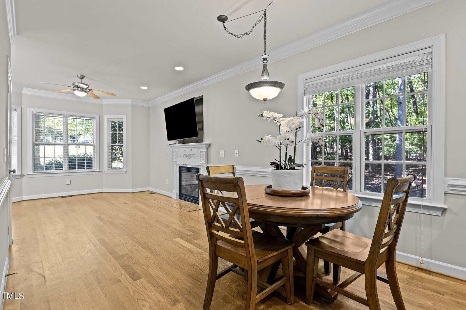 A dining area with a round wooden table and four chairs, set beside a window with a view of green foliage. The adjoining room features a fireplace and a wall-mounted TV. The space has hardwood floors and a ceiling fan with lights.