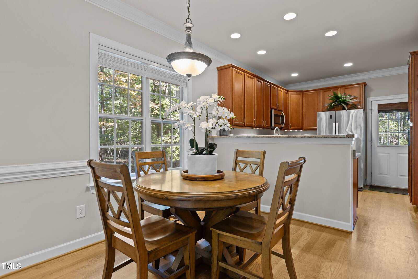 A kitchen with wooden cabinets and a dining area. A round wooden table with four chairs is centered under a hanging light fixture. A potted orchid decorates the table. Large windows let in natural light, highlighting the light wood flooring.