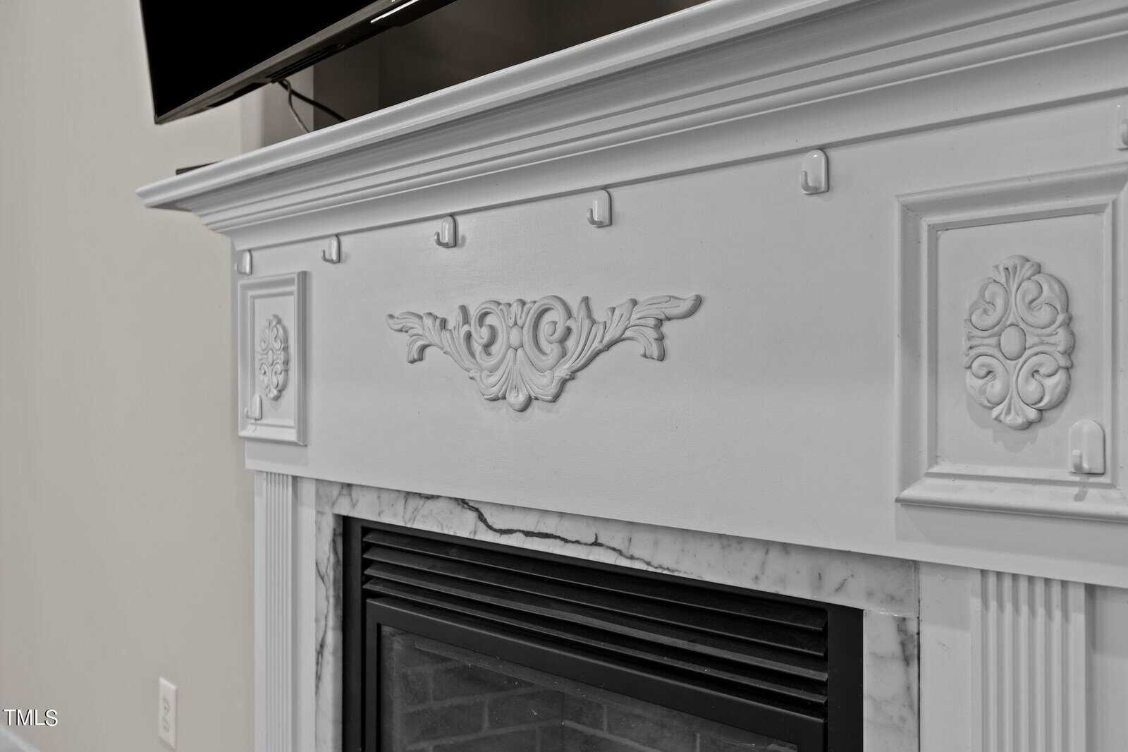 Close-up of an ornate white fireplace mantel with decorative carvings, including floral motifs and intricate patterns. The mantel features a classic design, and a television is partially visible above it.