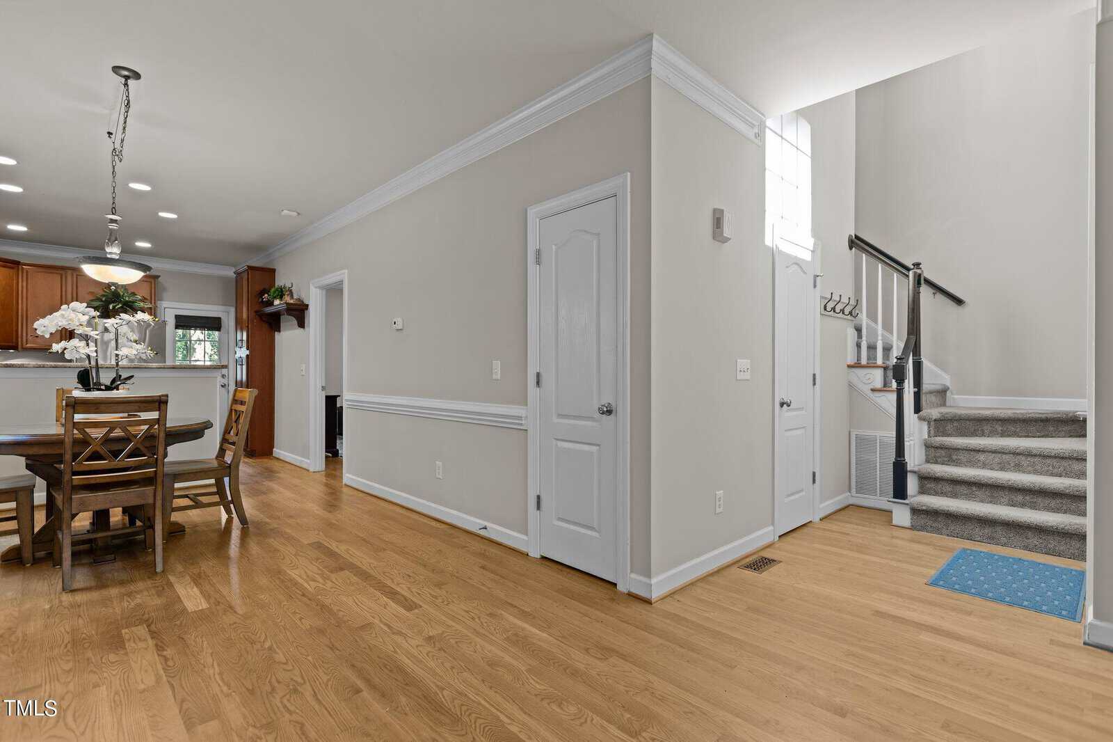 A modern interior with wooden floors features a dining area with a table and chairs on the left, and a kitchen with brown cabinets. A staircase with gray carpet leads upstairs on the right, next to white doors and light-colored walls.