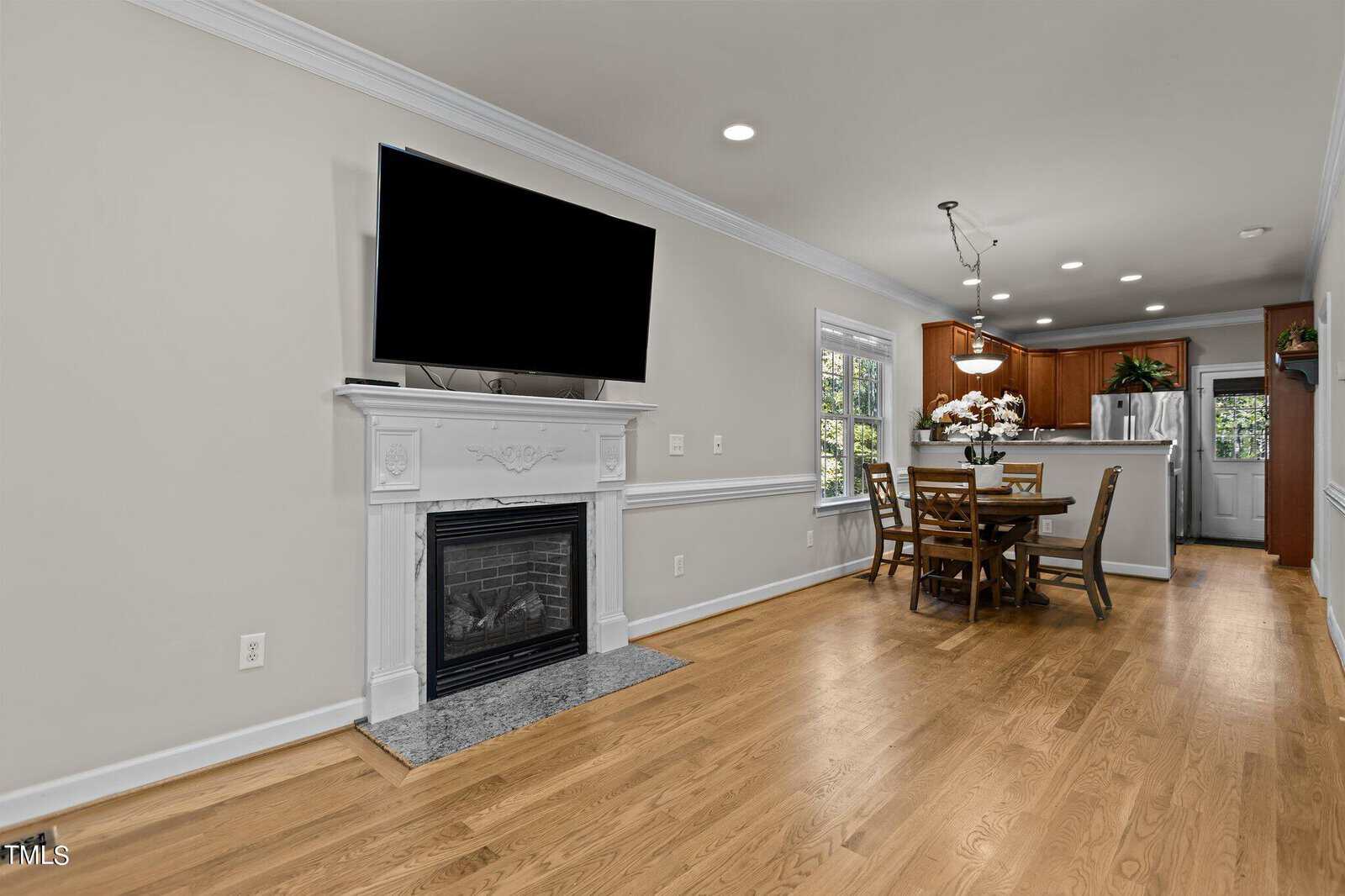 A modern living room features a mounted TV above a white fireplace, leading into a dining area with a wooden table and chairs. The room has hardwood floors and a view into a kitchen with wooden cabinets and a door to the outside.