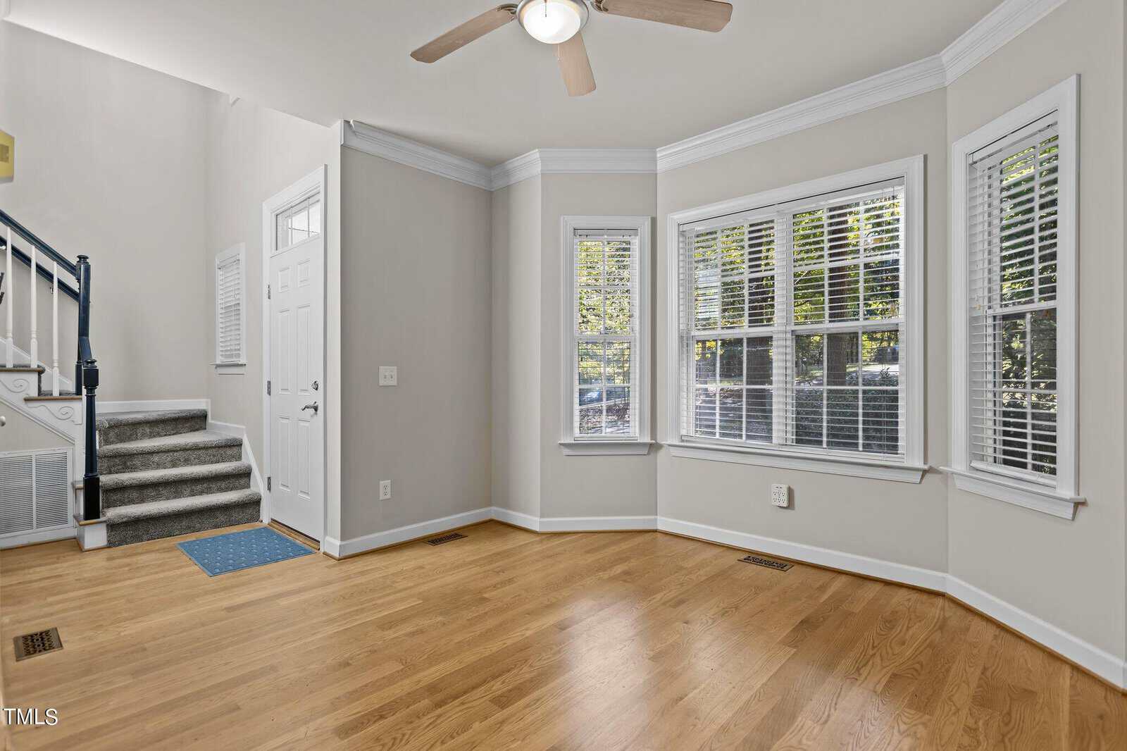 A bright, empty room with hardwood floors and three large windows with blinds. A ceiling fan hangs overhead. The white entrance door is next to a blue doormat, and there's a staircase with a black railing on the left.