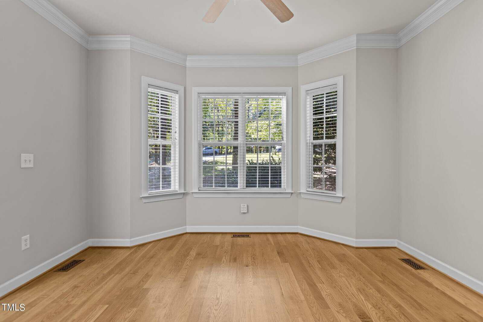 A bright, empty room with light gray walls and a wooden floor. Three large windows with white blinds allow natural light to fill the space. A light-colored ceiling fan is visible above.