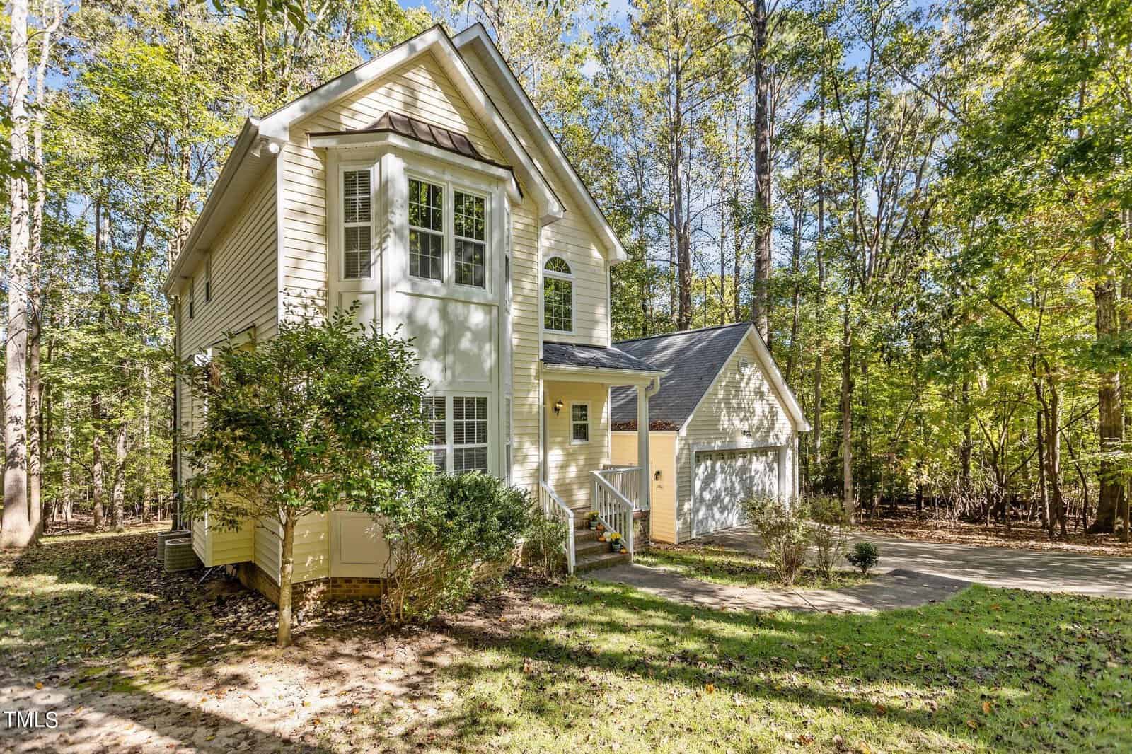 A two-story yellow house with a gray roof is surrounded by trees. The home has large windows and an attached garage. A pathway leads to the front entrance, and sunlight filters through the trees, casting shadows on the lawn.