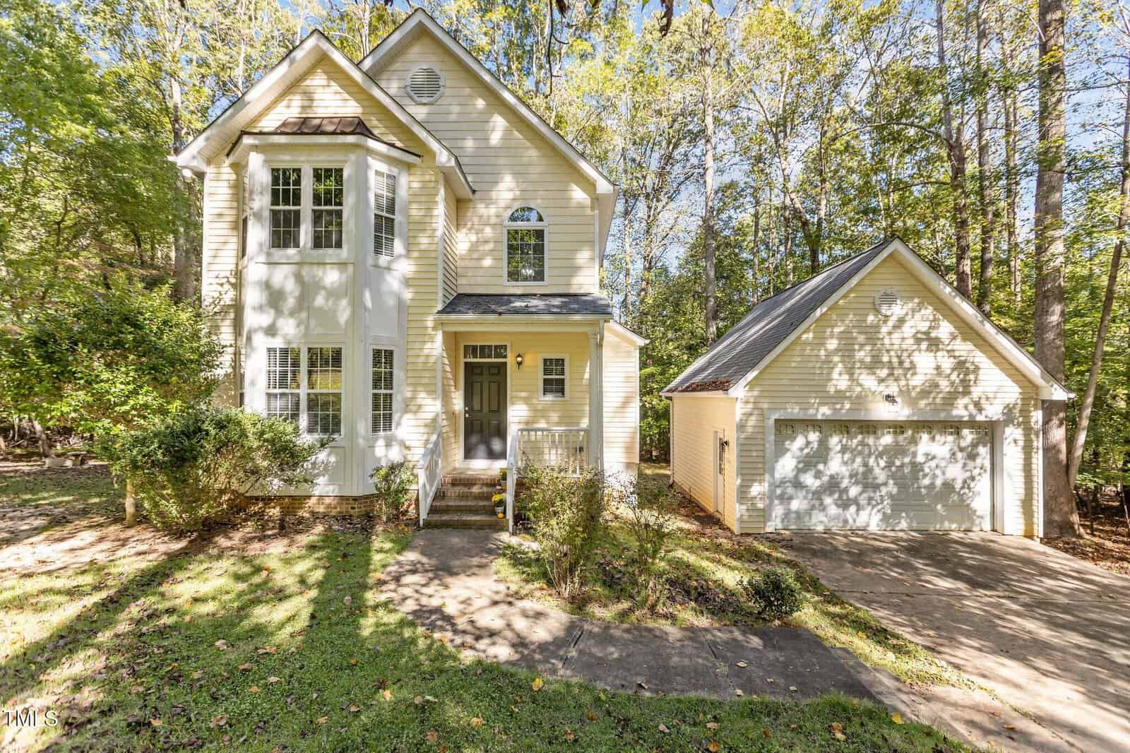 A charming two-story house with cream siding and a small front porch, nestled among tall trees at 3649 Wisteria Court. A detached two-car garage is to the right, and a path leads to the entrance. The surrounding yard in Wake Forest is adorned with scattered autumn leaves.