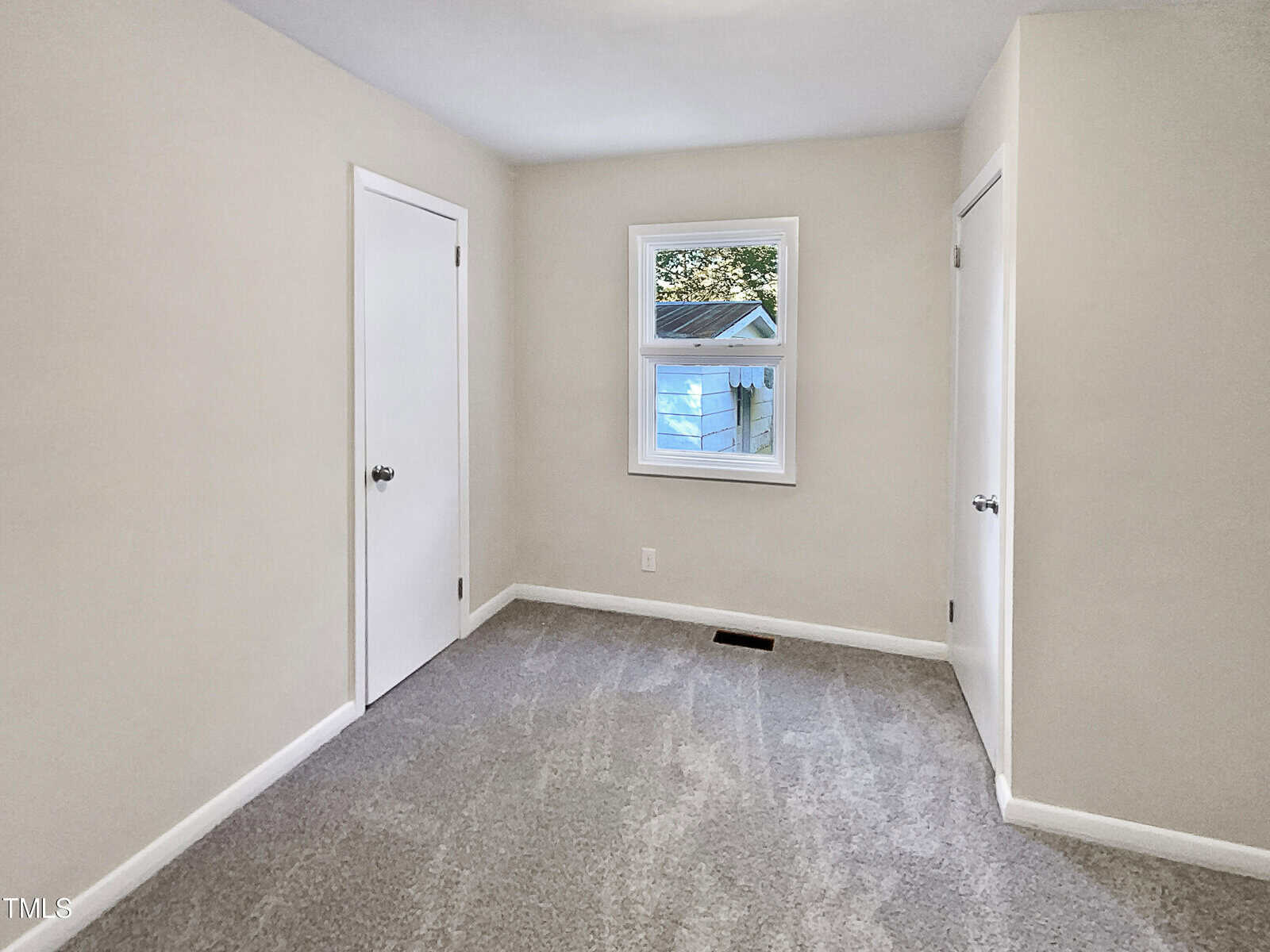 A small empty room with beige walls, gray carpet, a single window showing a house outside, and two white doors with silver handles.