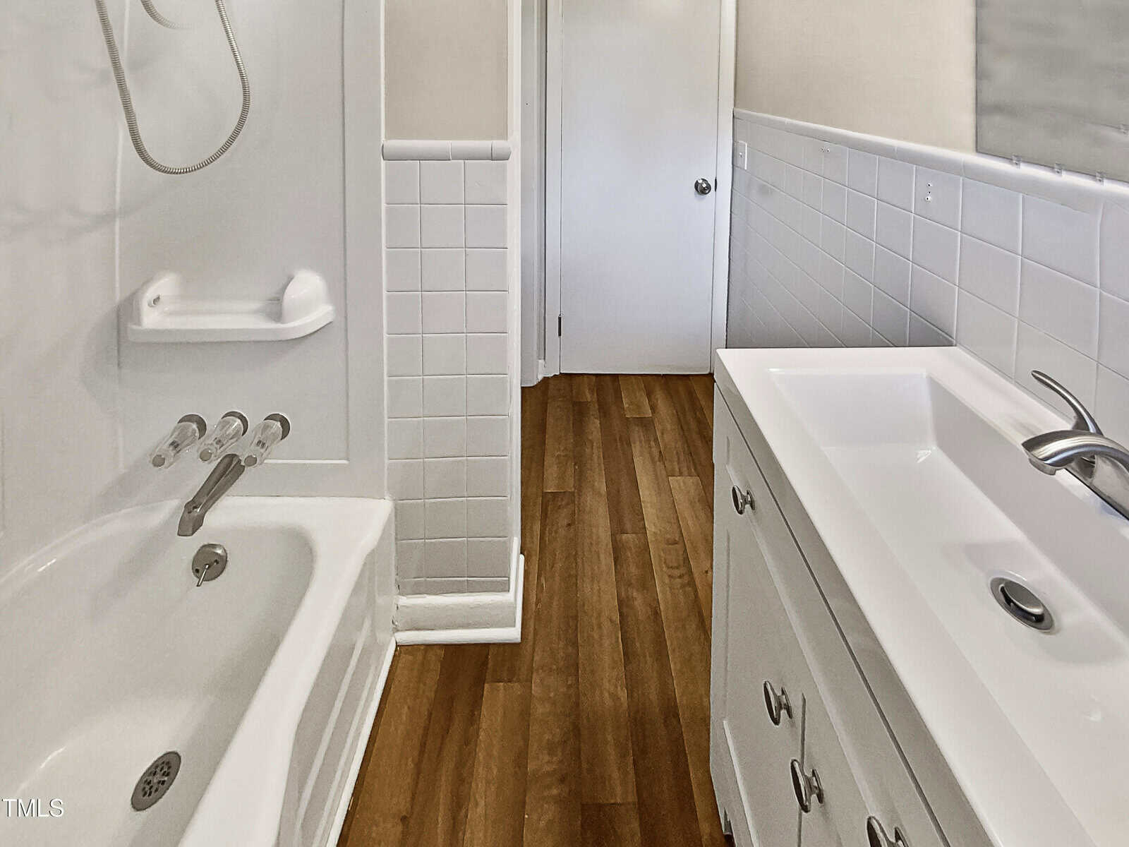 A small bathroom with a white tub-shower combo, white vanity with sink, silver faucet, tiled walls, and wood-style flooring. A door is at the far end of the narrow room.