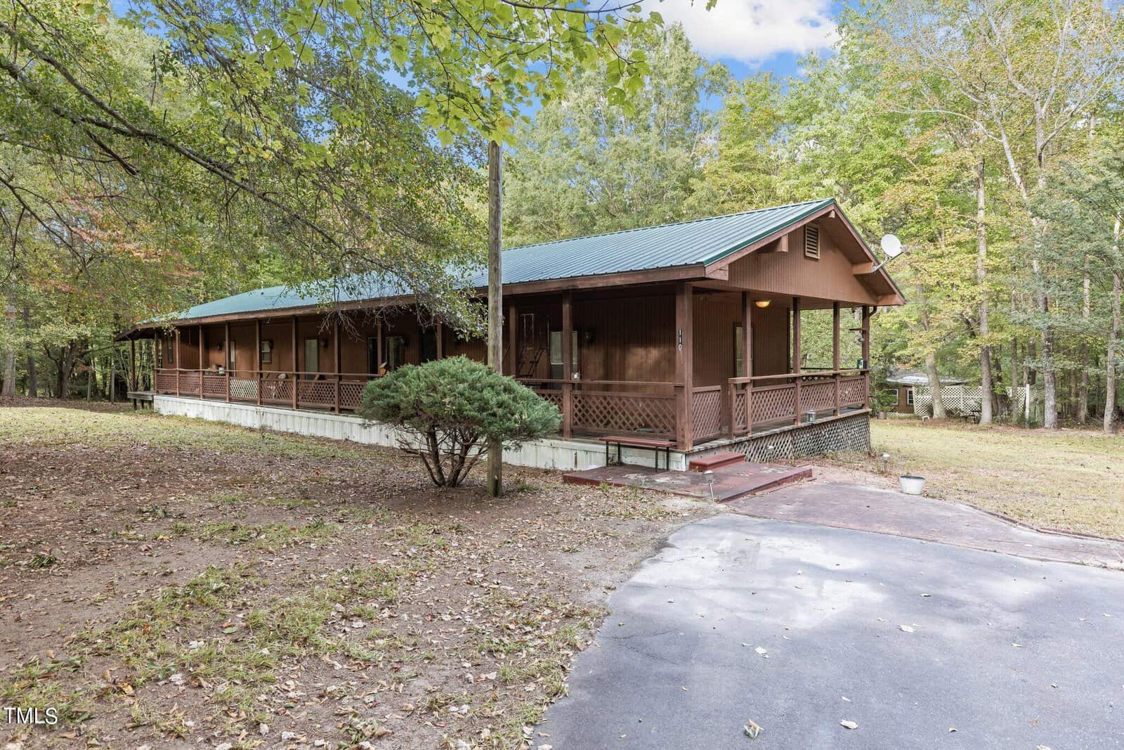 A single-story wooden house with a green metal roof, wraparound porch, and small shrub in front, surrounded by trees and a paved driveway leading up to the porch.
