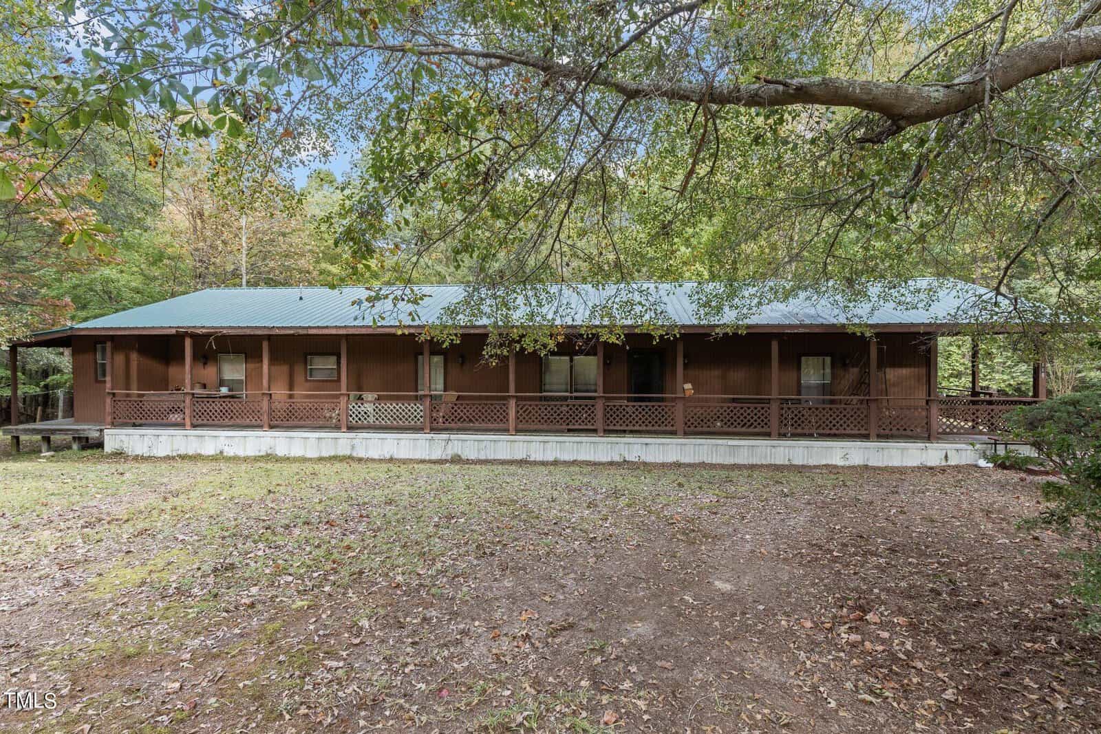 A long, single-story brown house with a green metal roof and a covered front porch, surrounded by trees and a yard with patchy grass and dirt.