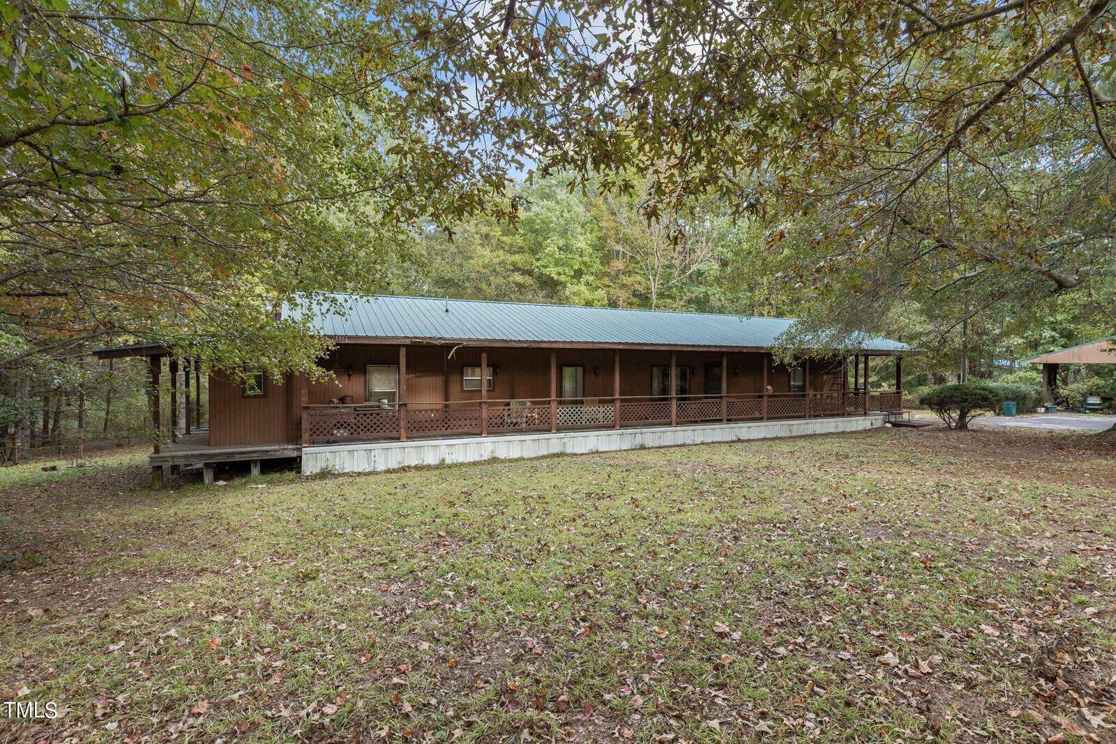 A single-story brown wooden house with a green metal roof and a long covered porch, surrounded by trees and a grassy yard covered with some fallen leaves.