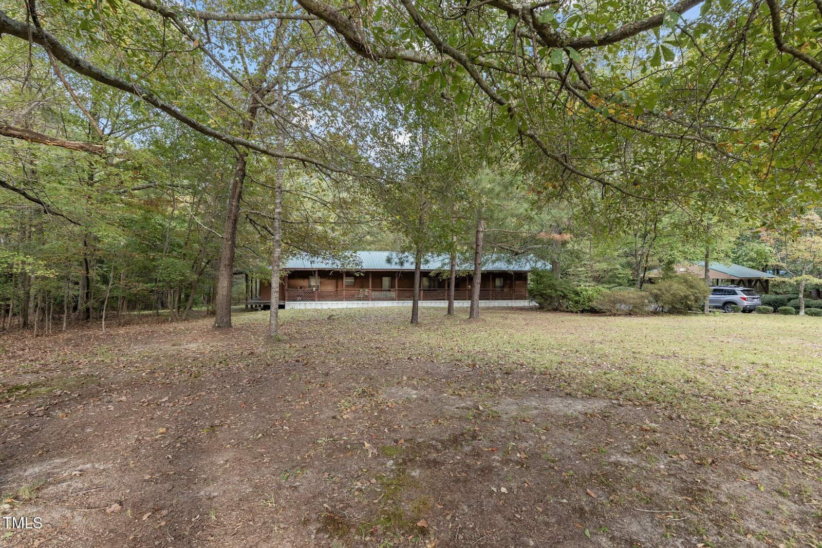 A single-story wooden house with a green metal roof is set back from the road, partially obscured by trees. The yard is mostly bare ground with some grass and scattered leaves. A vehicle is parked to the right.