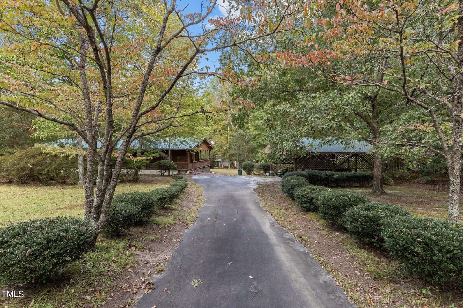 A paved driveway leads to a wooden house and a covered pavilion surrounded by trees and shrubs on a grassy lot. The scene is set during daytime in early autumn.