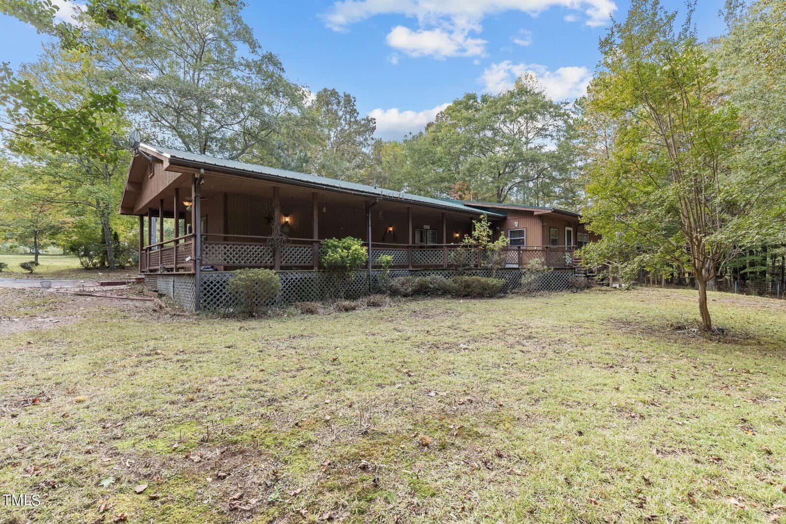 A single-story brown house with a large covered front porch and lattice skirting, surrounded by trees and a grassy yard under a partly cloudy sky.