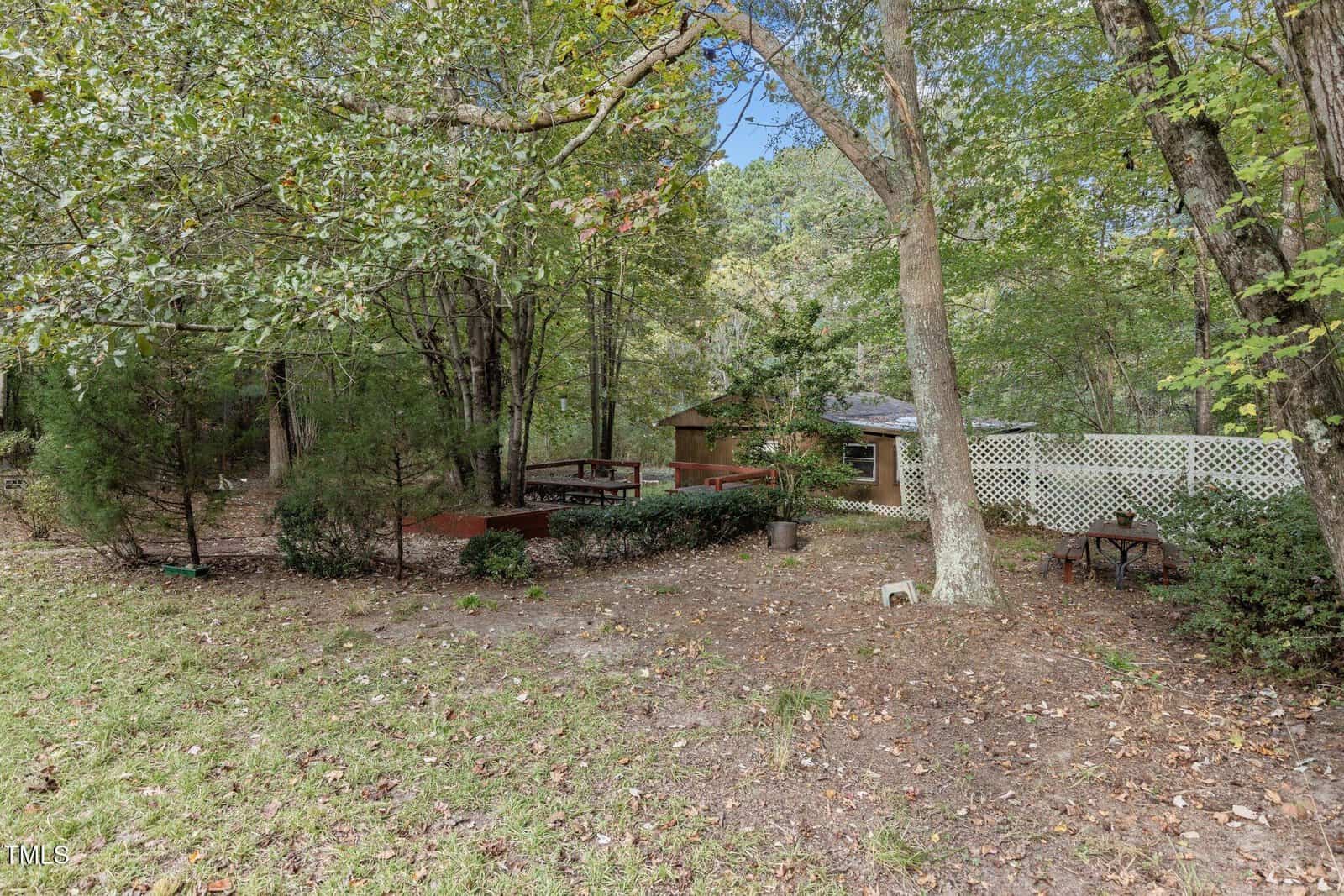 A backyard with large trees, a wooden deck, potted plants, a small shed, and a white lattice fence. Leaves cover the ground and patio furniture sits near the fence.