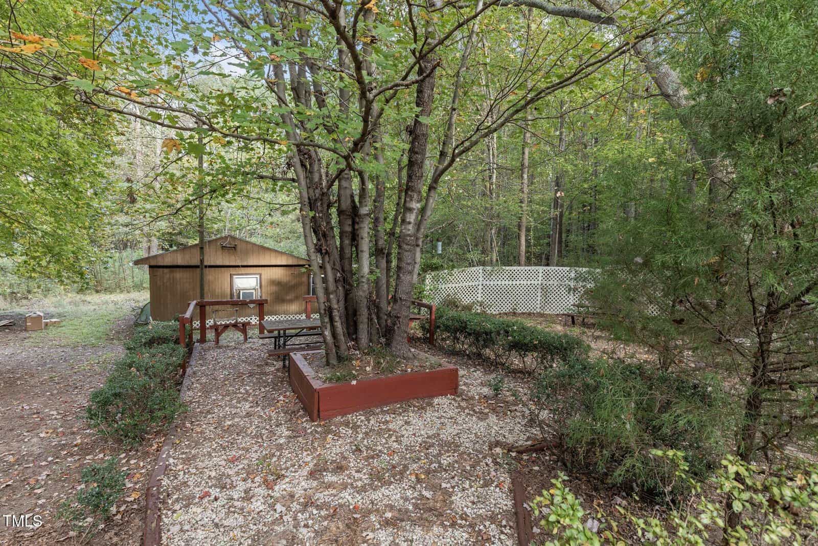 A small wooden shed sits behind a tree with a bench and raised flower bed in a gravel garden, surrounded by greenery and a white lattice fence in the background.