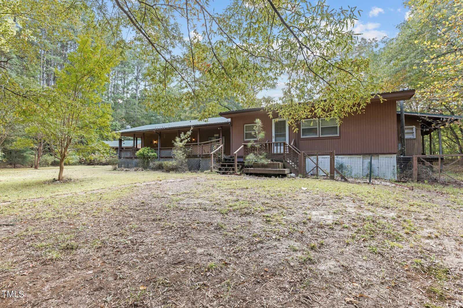 Single-story brown house with a porch, steps, and several windows, surrounded by trees and a grassy yard on a clear day.
