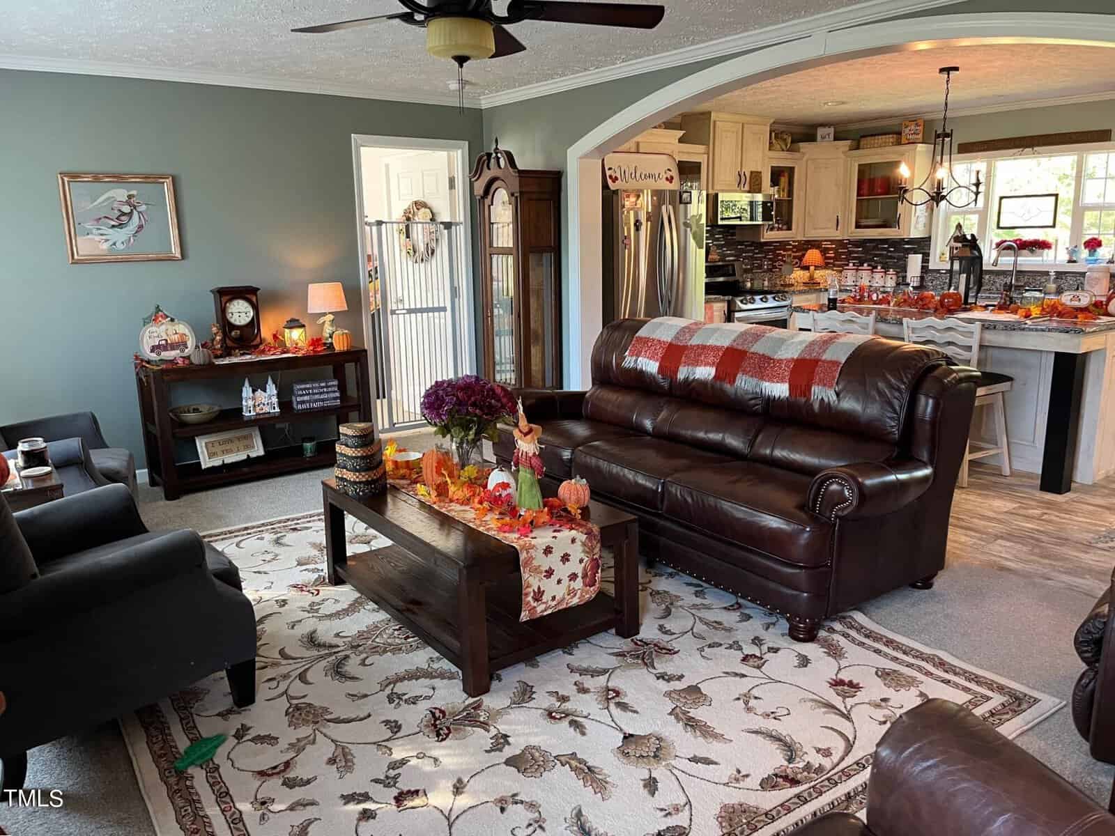 A cozy living room with leather sofas, a wooden coffee table decorated with autumn-themed items, and a grandfather clock. The kitchen is visible in the background, adorned with fall decorations. A ceiling fan hangs above a patterned rug.