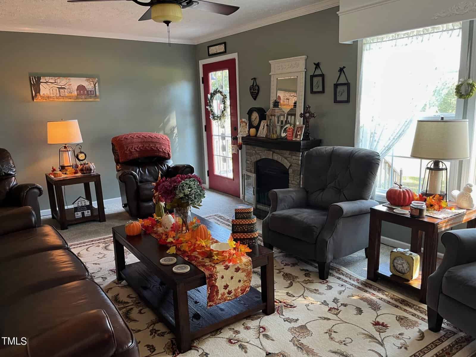 Cozy living room with autumn decor featuring a sofa, armchairs, and a fireplace. A coffee table displays pumpkins and leaves, while side tables hold lamps. A rug, ceiling fan, and wall art complete the warm, inviting atmosphere.