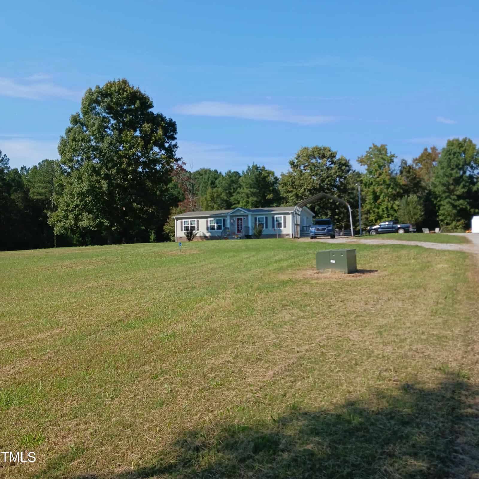 A single-story house sits in the distance surrounded by trees. A large grassy field extends in front of it, and a gravel driveway leads up to the home. A parked car is visible near the house under a clear blue sky.
