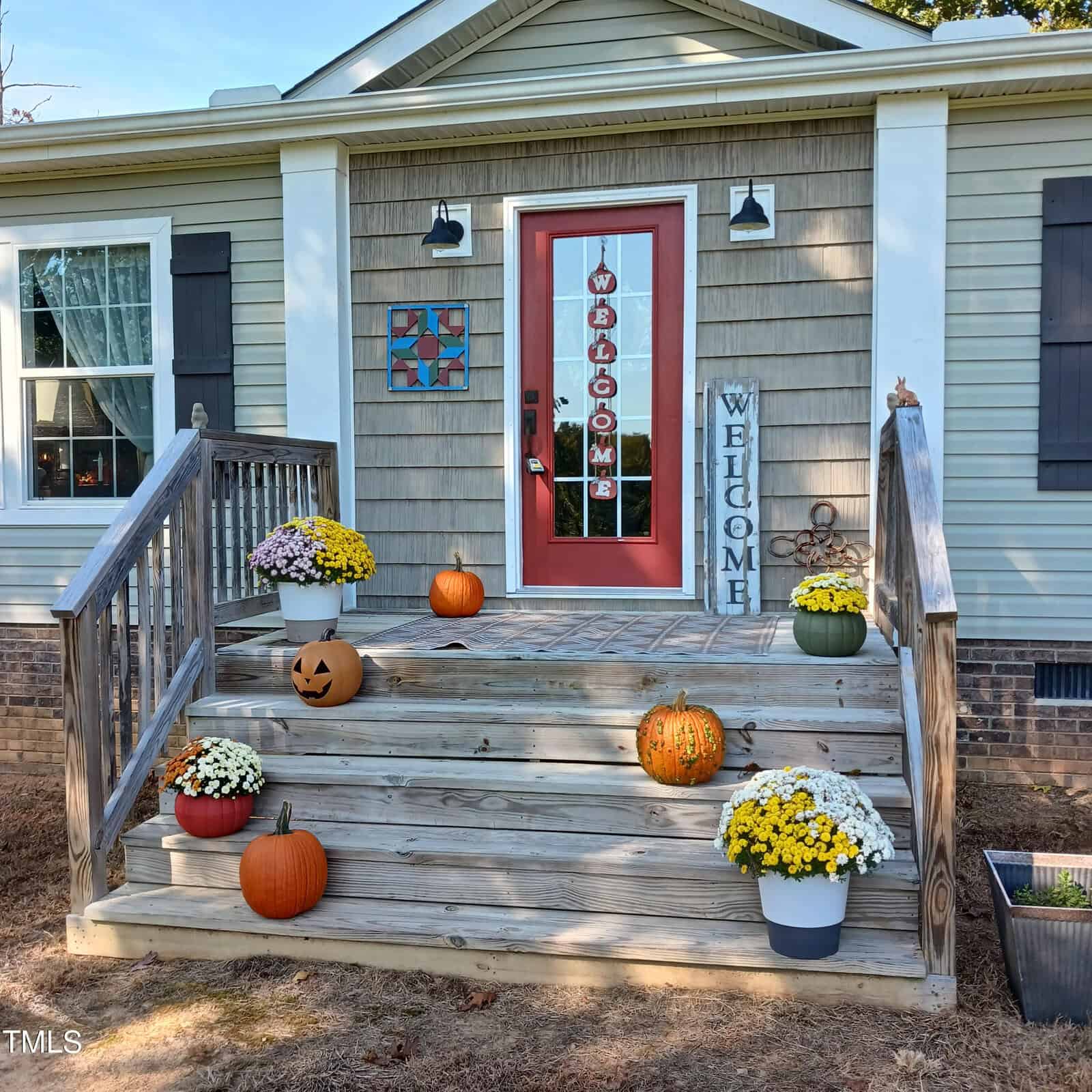 A cozy porch on 2679 Jack Yancey Road is decorated for fall with pumpkins, chrysanthemum flowers, and a "Welcome" sign on the steps. The front door has a red frame with a vertical "Welcome" sign, and the exterior is light gray with white trim, reflecting the charming Bullock style.