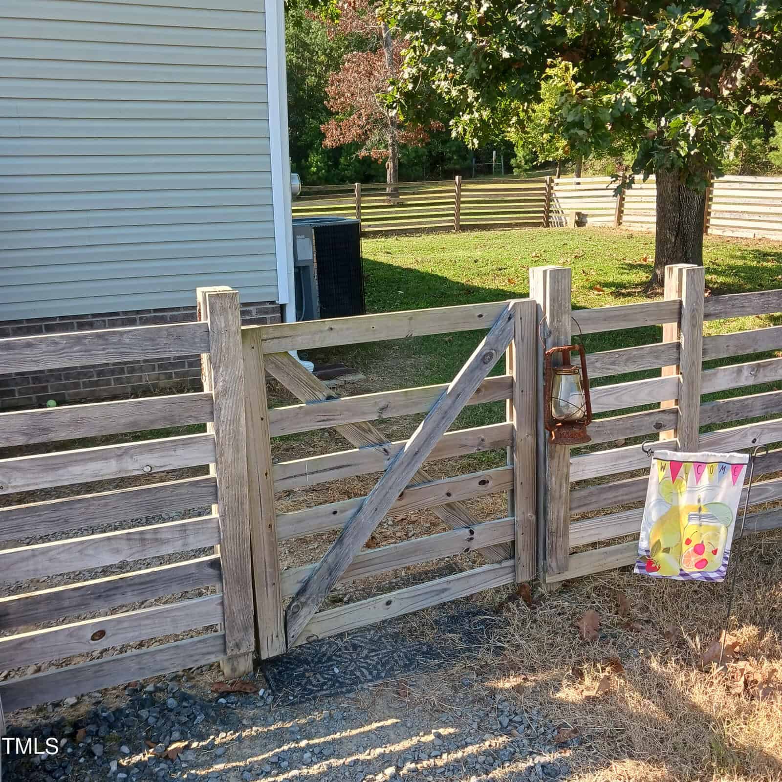A wooden fence gate surrounded by a yard with a tree. A colorful flag with lemon designs hangs on the fence, and an old lantern is attached to the post. A house and an air conditioning unit are visible in the background.