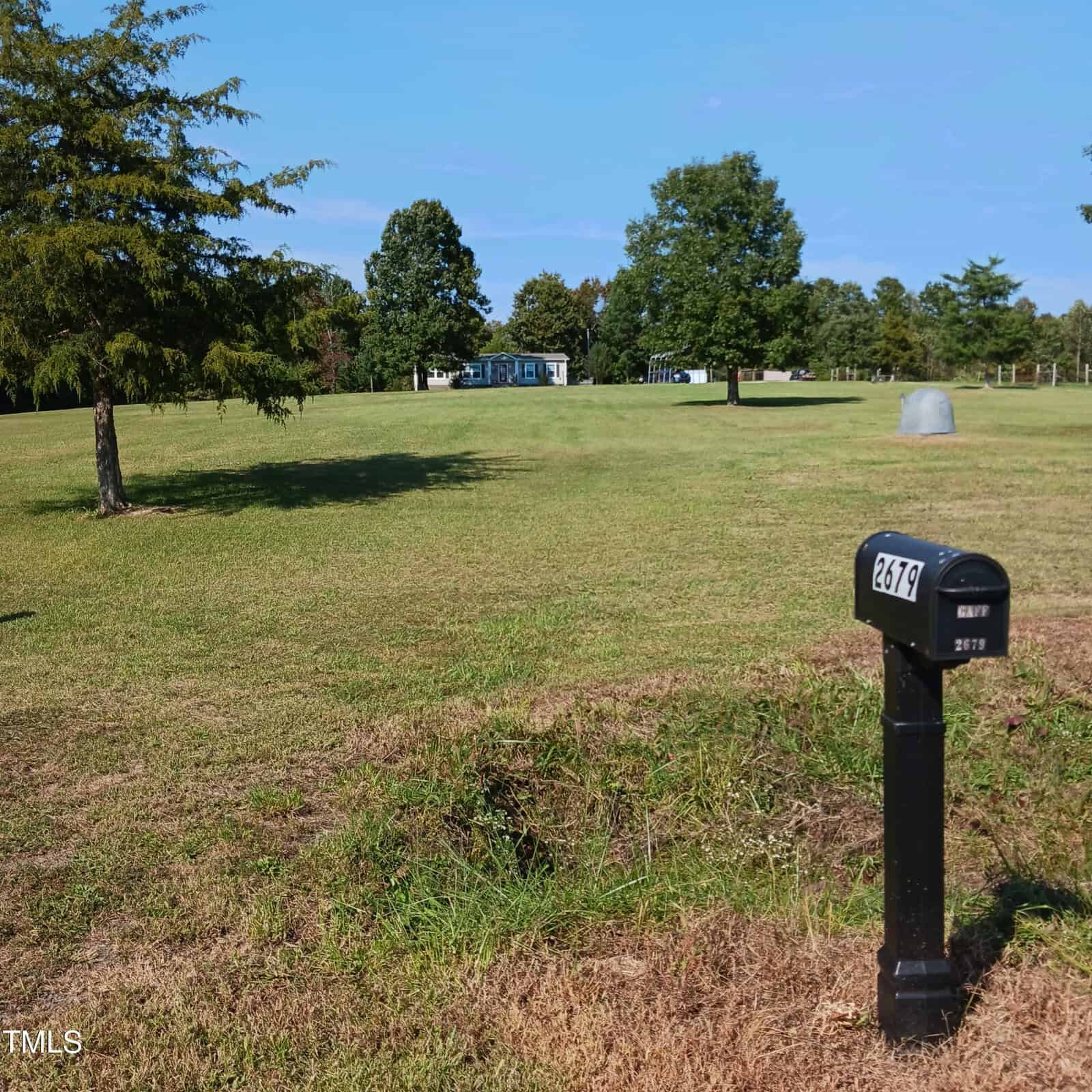 A black mailbox with the number 2679 stands in a grassy field under a clear blue sky. Trees and a distant structure are visible in the background.